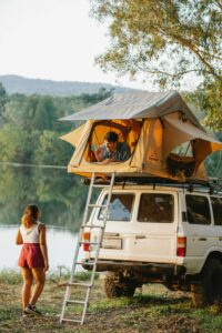 Couple camping by a lake with a rooftop tent mounted on a white Toyota Land Cruiser SUV. A man is seen smiling and waving from the Everest rooftop tent while a woman in athletic wear stands on the ground beside the vehicle. The setting is a serene lakeside surrounded by lush trees and distant hills, captured during golden hour.