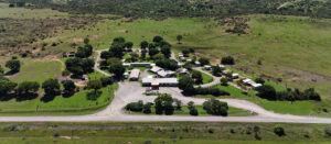Aerial photograph of a rural caravan park and campground nestled in a wide expanse of green countryside, with small cabins, trailers, and administrative buildings arranged along winding gravel roads. The site is shaded by scattered trees and bordered by a main road in the foreground. Rolling hills and distant mountain ranges dominate the background under a clear blue sky, suggesting a remote, peaceful setting ideal for eco-tourism, camping, and nature retreats. Keywords: rural caravan park, campground aerial view, countryside accommodation, nature getaway, eco-tourism site, cabins and trailers, scenic landscape, mountain backdrop.
