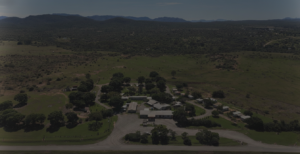 Aerial photograph of a rural caravan park and campground nestled in a wide expanse of green countryside, with small cabins, trailers, and administrative buildings arranged along winding gravel roads. The site is shaded by scattered trees and bordered by a main road in the foreground. Rolling hills and distant mountain ranges dominate the background under a clear blue sky, suggesting a remote, peaceful setting ideal for eco-tourism, camping, and nature retreats. Keywords: rural caravan park, campground aerial view, countryside accommodation, nature getaway, eco-tourism site, cabins and trailers, scenic landscape, mountain backdrop.