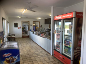 Interior view of a modest rural café and convenience store featuring a tiled floor, ceiling fans, and a long counter with food service equipment. On the right, Coca-Cola branded refrigerated display cases are stocked with soft drinks, water, and juice bottles. A freezer displaying Peters ice cream is positioned on the left. Behind the counter, a small shop area contains shelves and coolers with snacks and grocery items. Handwritten chalkboard menus and printed signs are mounted on the wall, contributing to a casual, local-store atmosphere. Keywords: rural café interior, convenience store, Coca-Cola fridge, ice cream freezer, snack bar, small business, country shop, takeaway food counter, local grocery.