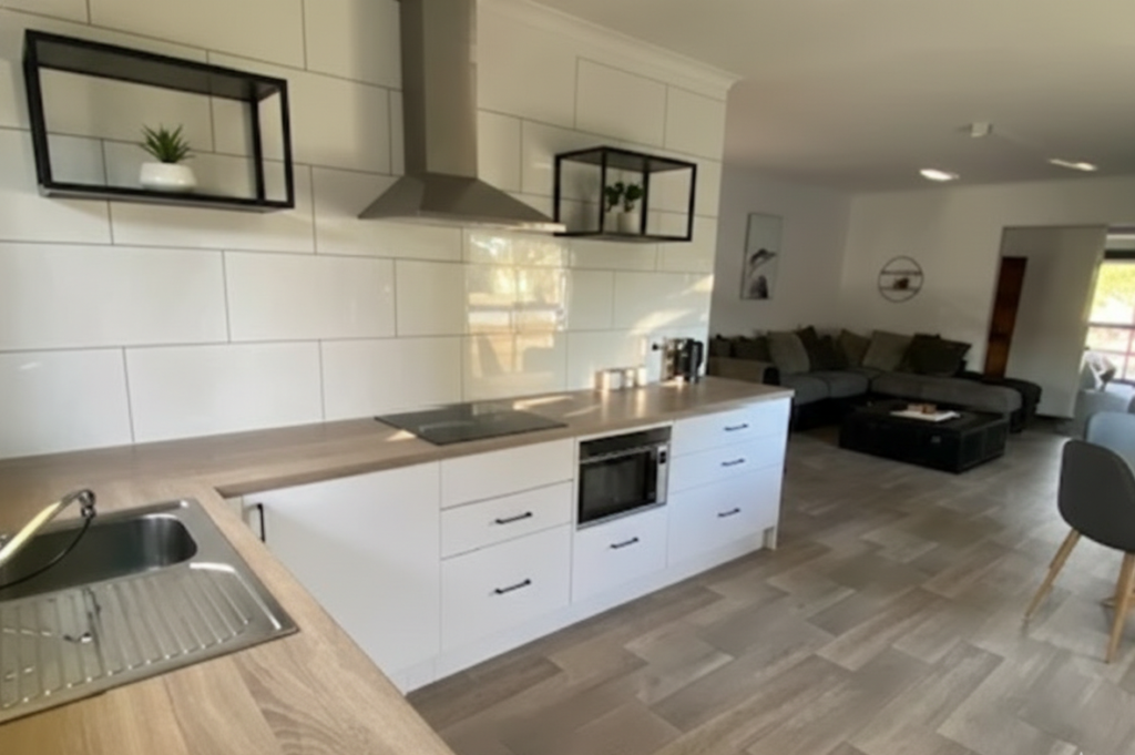 Contemporary open-plan kitchen and living space featuring white cabinetry with integrated appliances, including a built-in microwave and sleek rangehood. The kitchen has light wood countertops, a stainless steel sink, and white subway tile backsplash. Floating black metal-framed shelves with small potted plants add a decorative touch. The flooring is light wood-look tile that extends into the adjoining living room, which contains a dark sectional sofa and modern wall décor. Natural light filters through the space, enhancing the clean, minimalist design. Keywords: modern kitchen interior, open-plan living, minimalist home design, white cabinetry, wood countertops, tiled floor, contemporary apartment, kitchen and living combo.